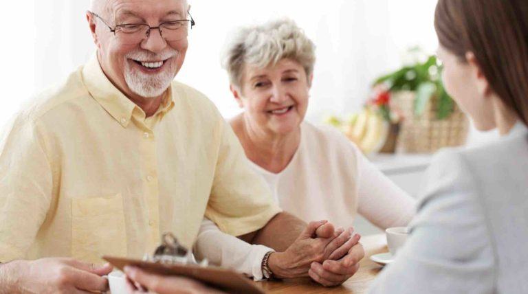 Älteres Ehepaar sitzt am Tisch und lächelt, halten Händchen, andere Frau zeigt dem Ehepaar Dokument